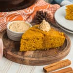 A slice of pumpkin cornbread that is orange in color, on a wooden plate topped with maple butter. With a side dish of maple butter, a pumpkin towel, and 2 cinnamon sticks in the foreground.