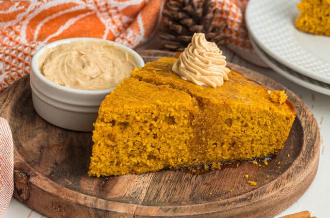 A slice of pumpkin cornbread that is orange in color, on a wooden plate topped with maple butter. With a side dish of maple butter, a pumpkin towel, and 2 cinnamon sticks in the foreground.