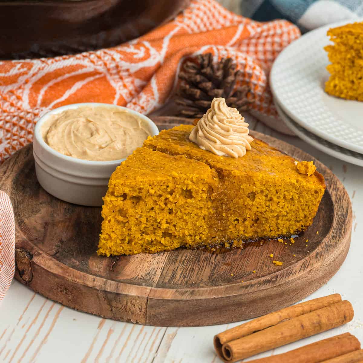 A slice of pumpkin cornbread that is orange in color, on a wooden plate topped with maple butter. With a side dish of maple butter, a pumpkin towel, and 2 cinnamon sticks in the foreground. 