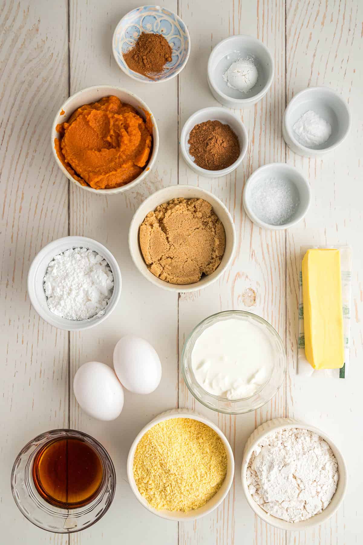 Ingredients for pumpkin cornbread in small white bowls on a white wood surface, top view. 