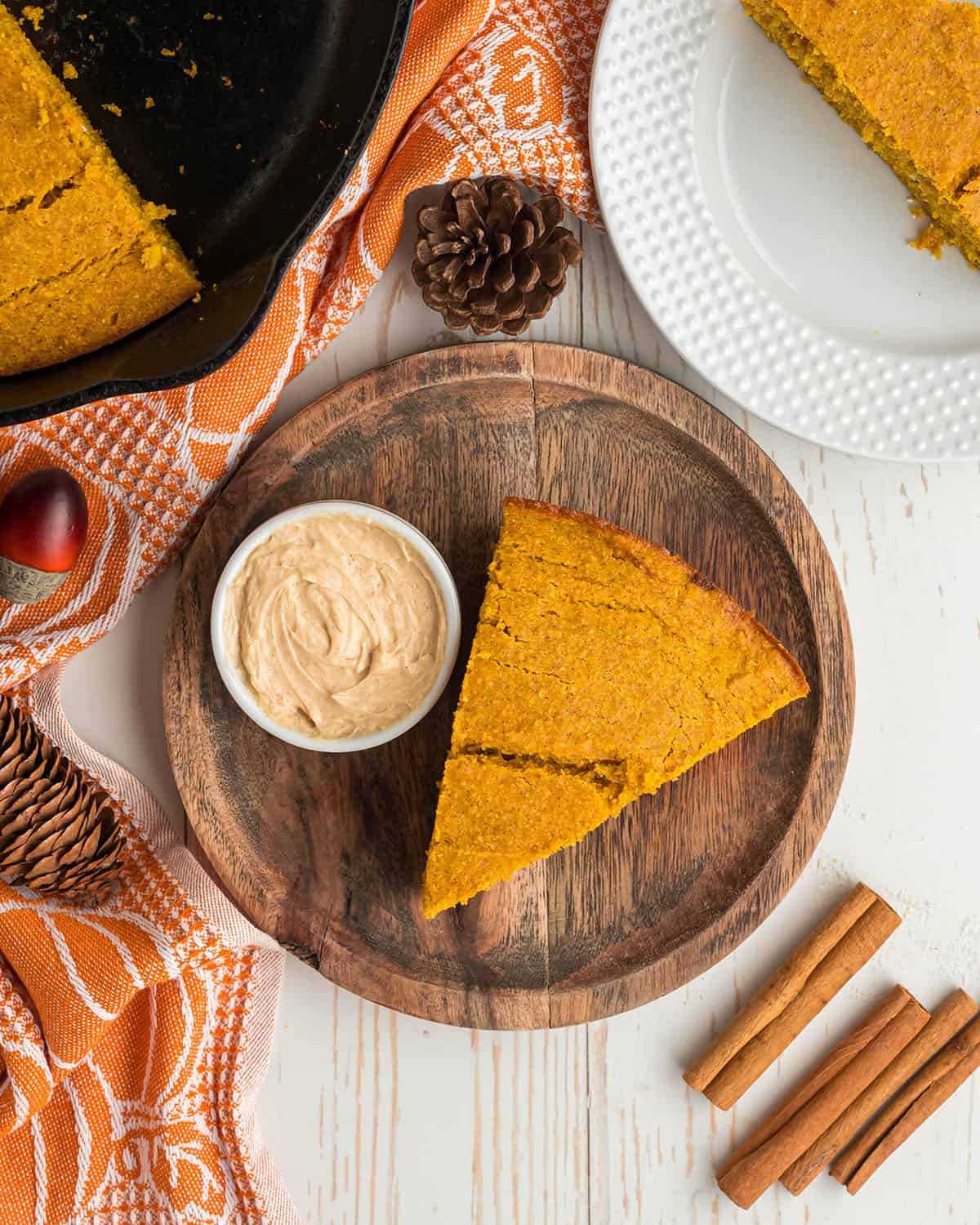 A slice of pumpkin cornbread on a wood plate surrounded by a small dish of maple butter, cinnamon sticks, and an orange towel.