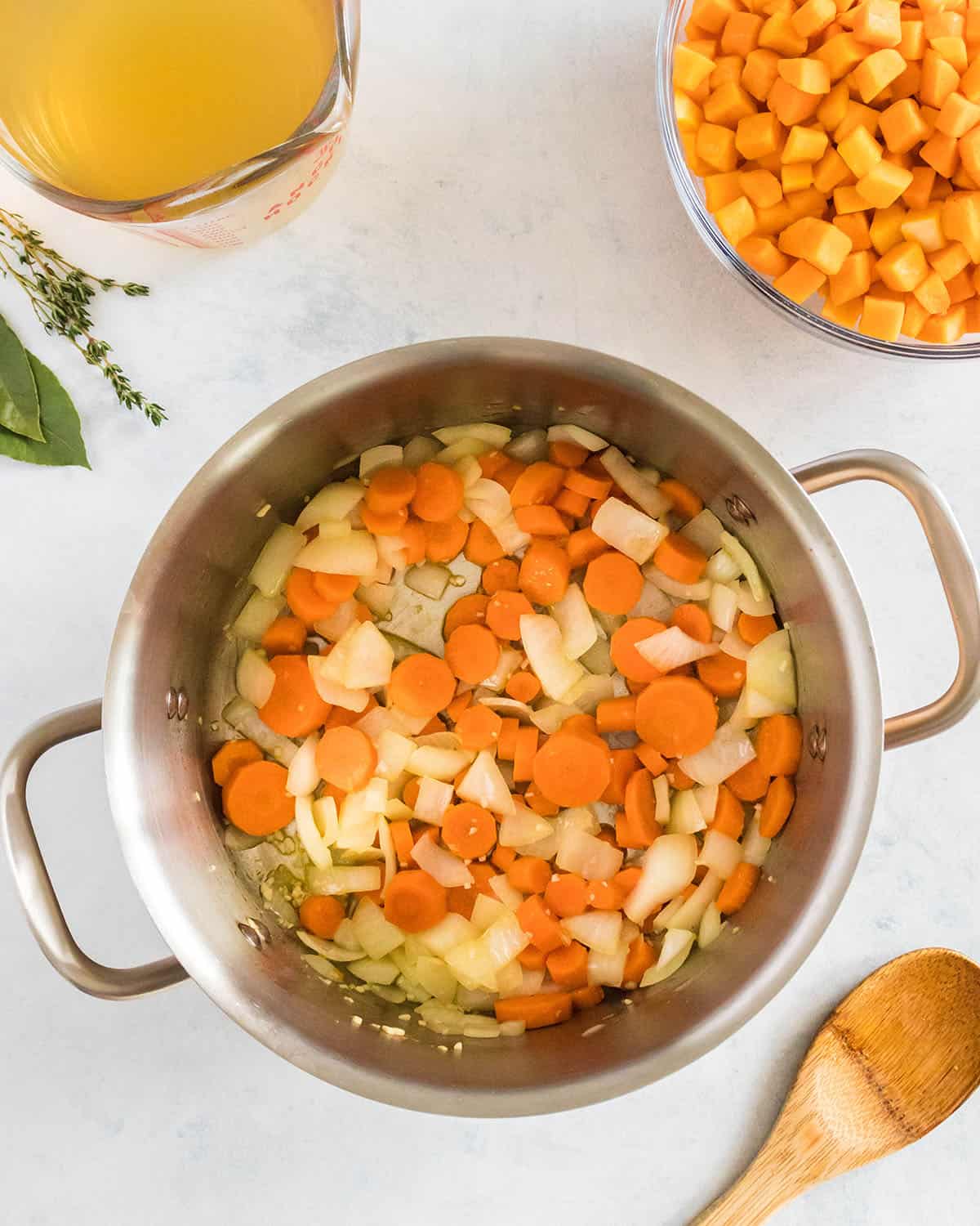 Vegetables cooking in a large soup pot, with a bowl of butternut squash cubes and a bowl of broth to the side. On a white surface, top view. 