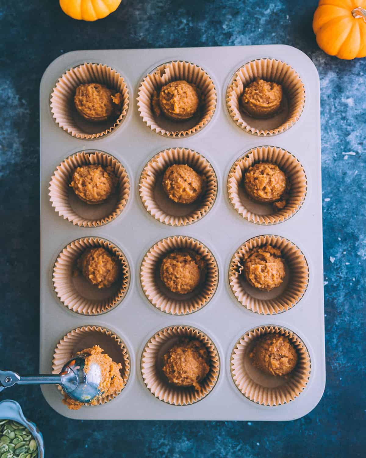 A muffin tin lined with paper cups with scoops of pumpkin cup batter in it. 