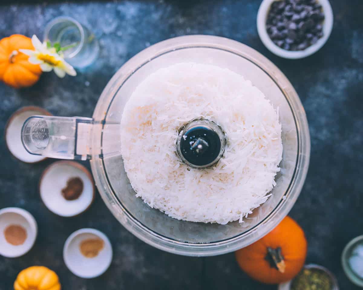 Shredded coconut in a food processor, surrounded by bowls of spices and fresh pumpkins. Top view. 