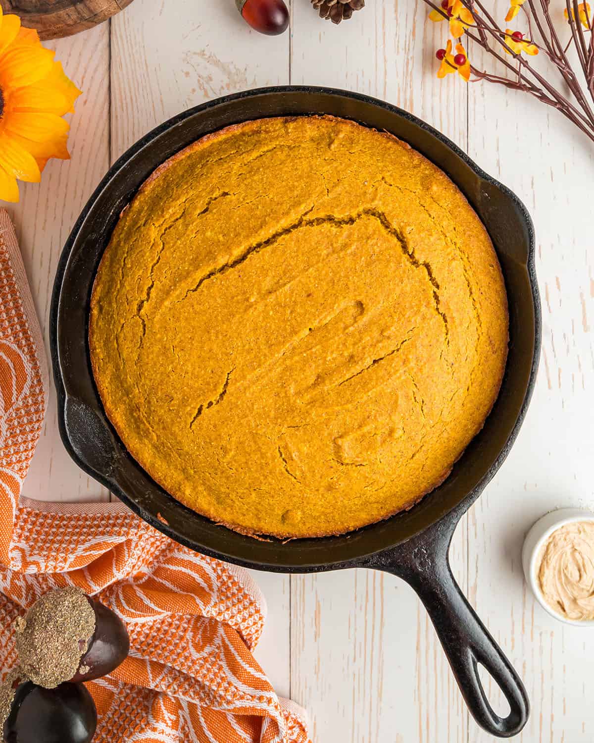 Baked pumpkin cornbread in a skillet, cooling and surrounded by an orange and white towel and fall flowers. 