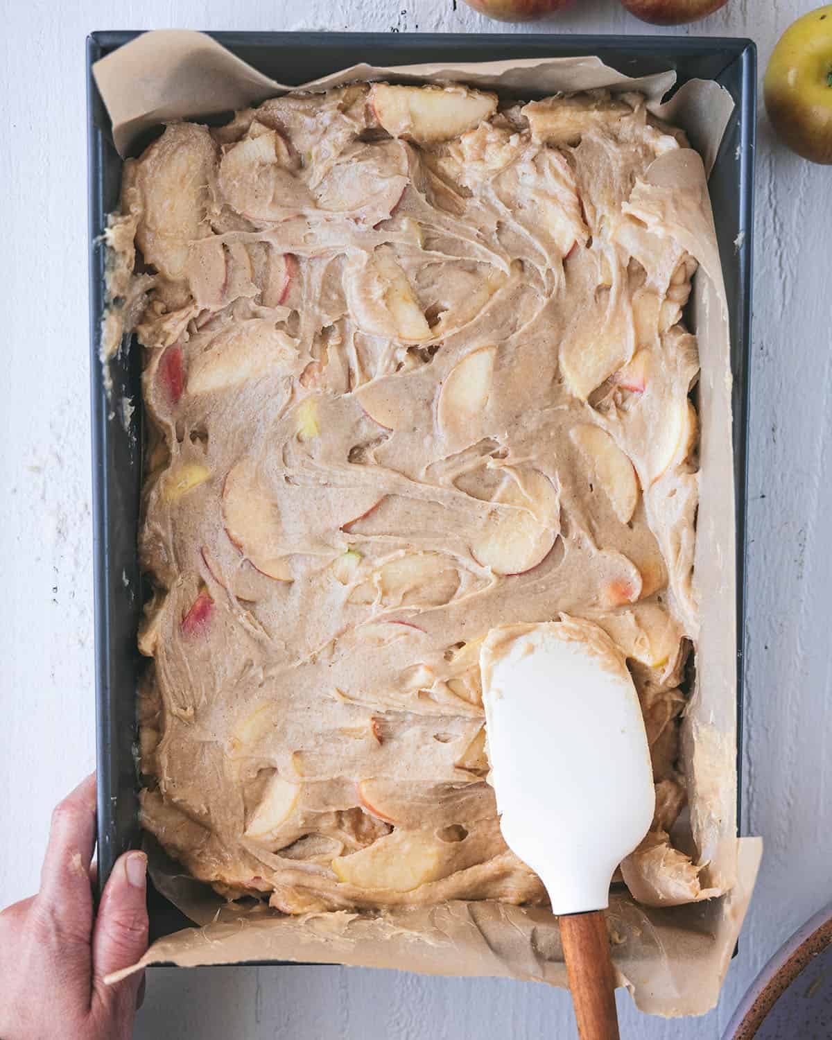 A rectangle pan with apple cake batter in it, being spread with a white rubber spoon. 