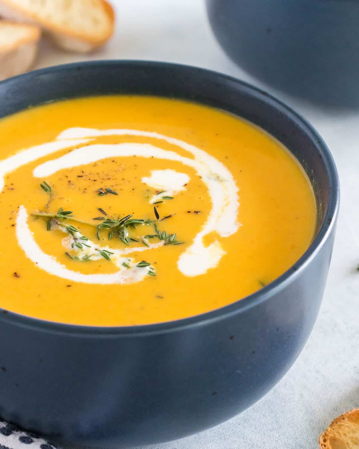 A bowl of butternut squash soup topped with a drizzle of cream and spices, surrounded by bread slices, fresh thyme, and a napkin and spoon. 