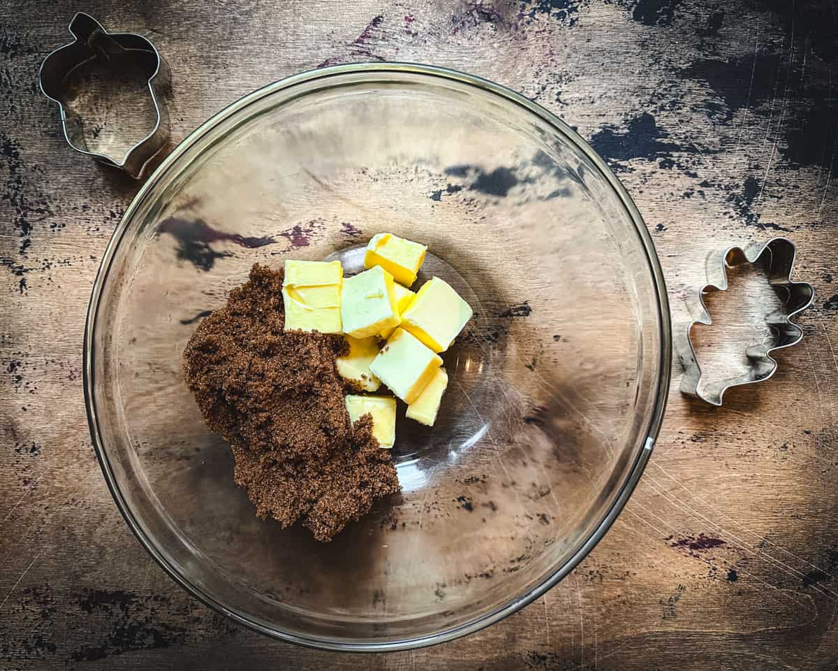 Butter and brown sugar in a mixing bowl, on a wood surface surrounded by cookie cutters. Top view. 