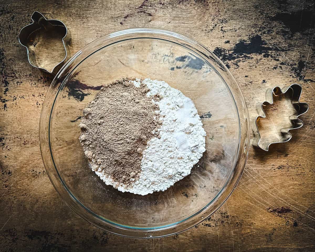 Acorn and regular flour mixing with dry ingredients in a mixing bowl, on a wood surface surrounded by cookie cutters on a wood surface. Top view.