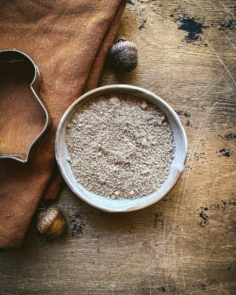 A bowl of acorn flour on a wood surface surrounded by a brown cloth, felt acorns, and an acorn cookie cutter shape.