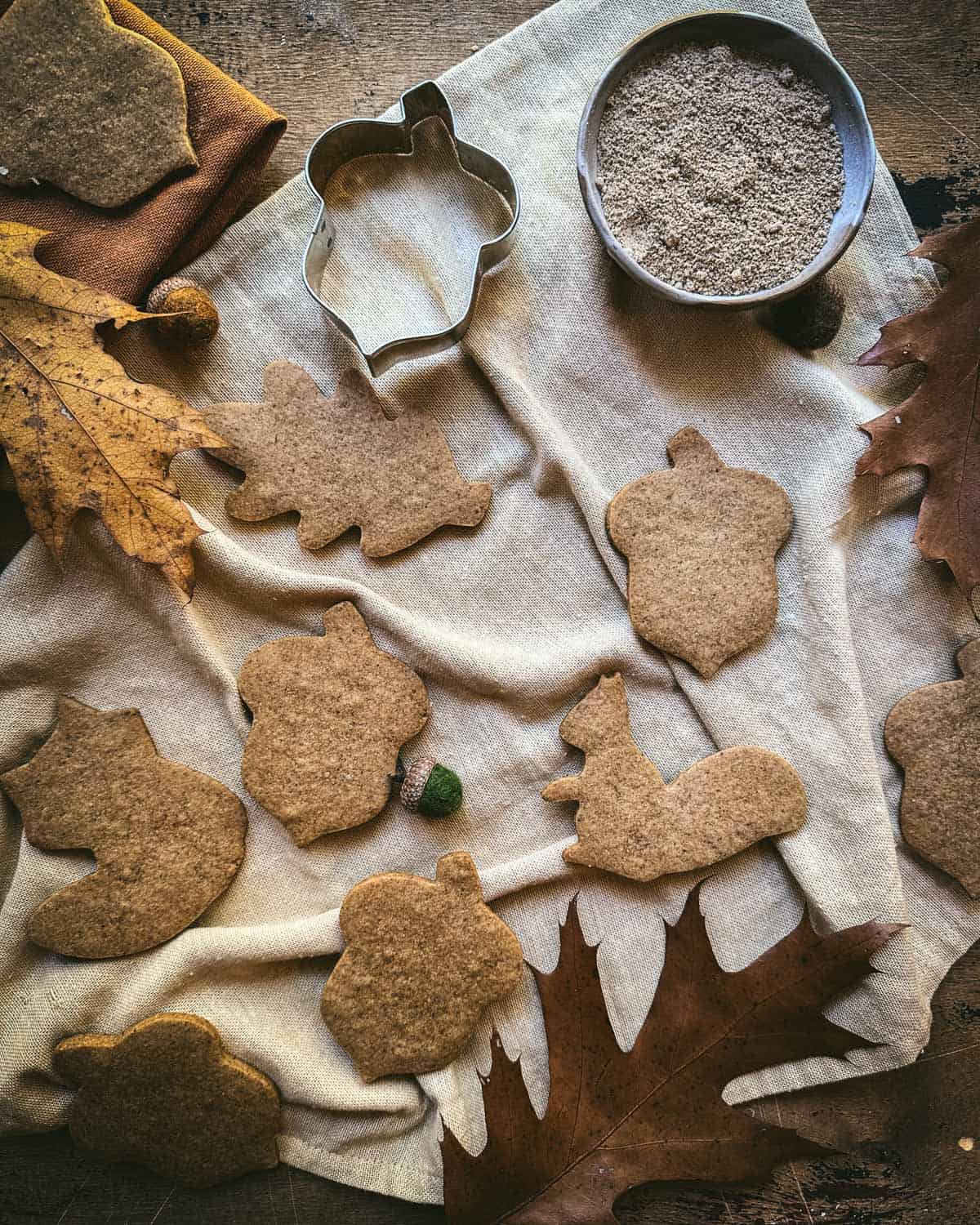 Acorn flour cookies in fall shapes on a white fabric, surrounded by an acorn cookie cutter, fall leaves, and a bowl of acorn flower. 