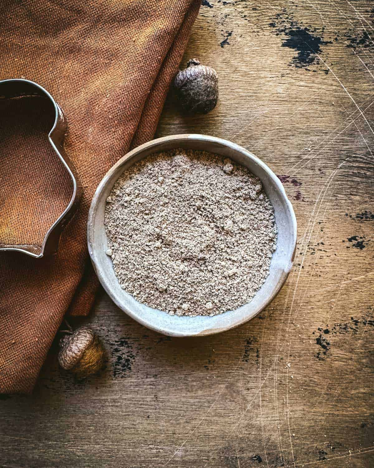 A bowl of acorn flour on a wood surface surrounded by a brown cloth, felt acorns, and an acorn cookie cutter shape. 