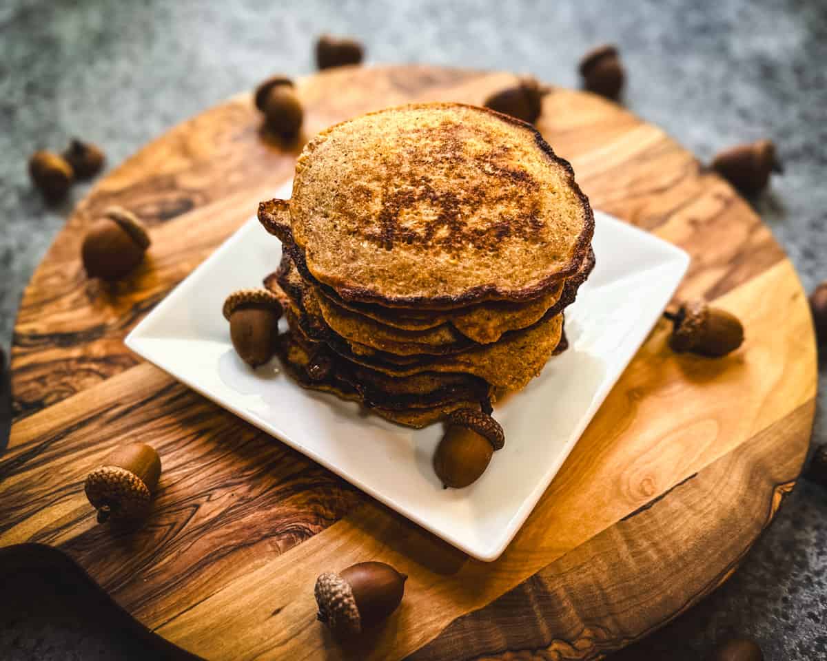 A white square plate with a stack of acorn pancakes on it, on a wood cutting board surrounded by acorns. 