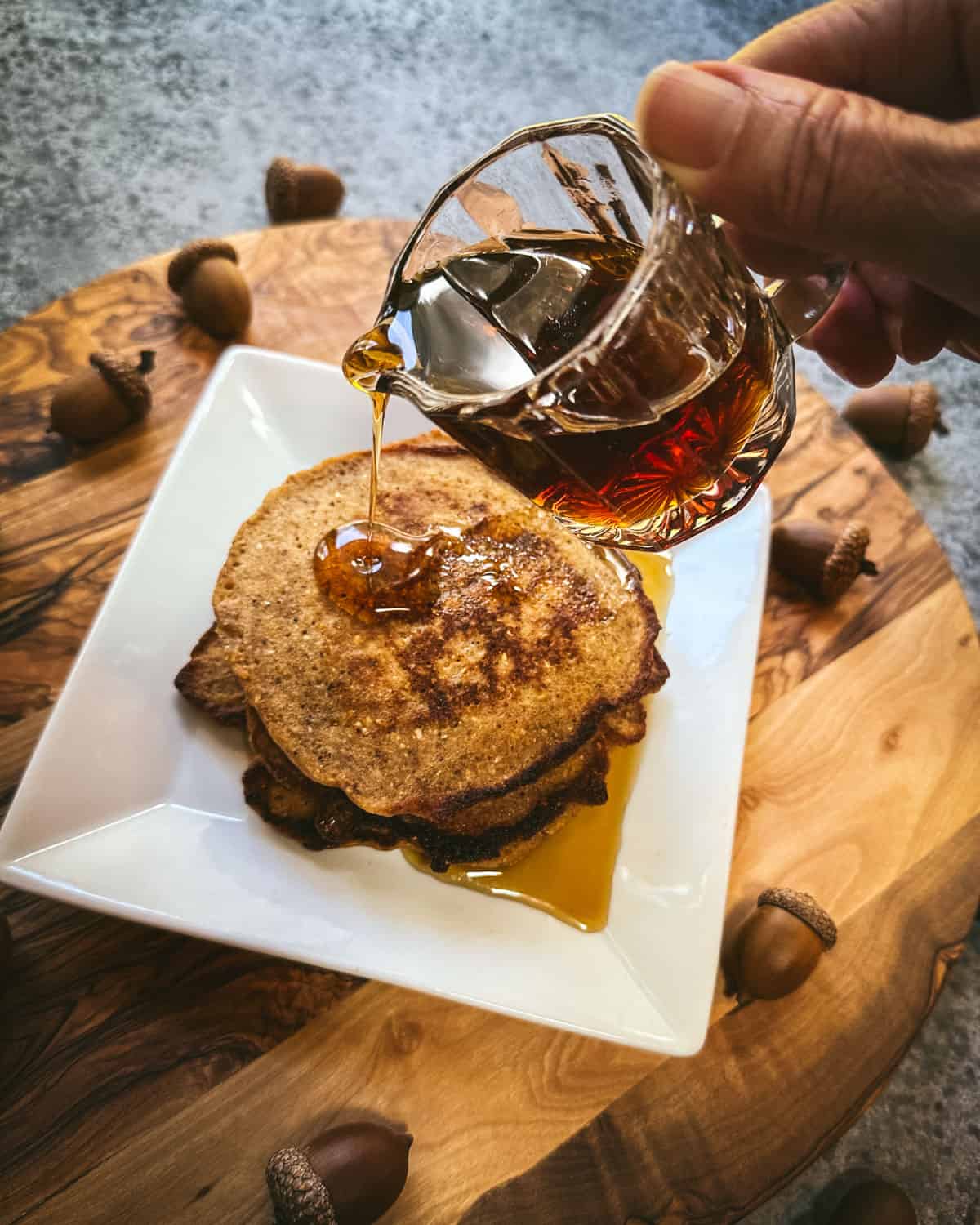 Acorn pancakes on a square white plate with syrup pouring onto it, on a wood cutting board surrounded by acorns. 