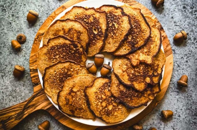 A plate with acorn pancakes layered in a circle, on a round wood cutting board, on a gray countertop surrounded by acorns.