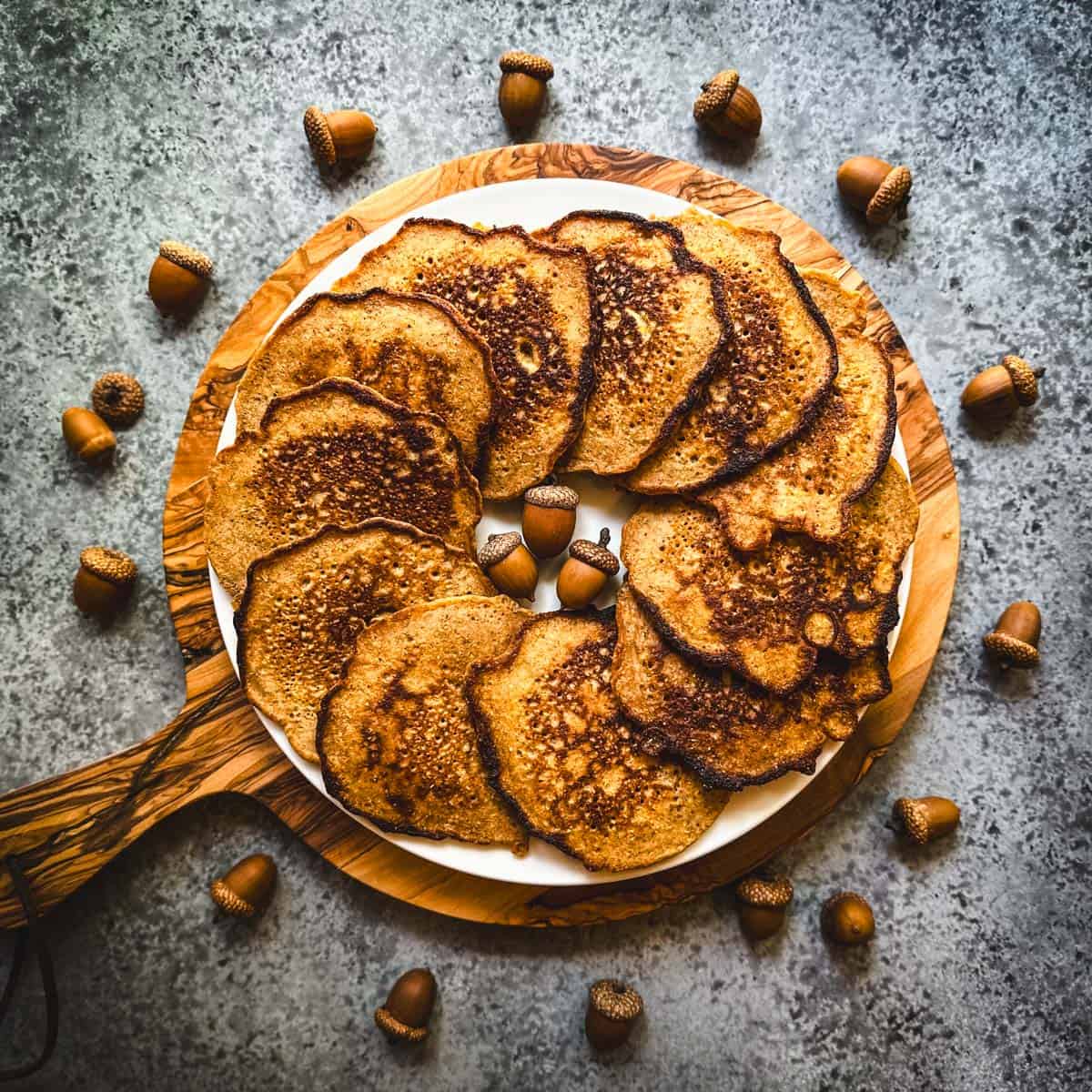 A plate with acorn pancakes layered in a circle, on a round wood cutting board, on a gray countertop surrounded by acorns. 
