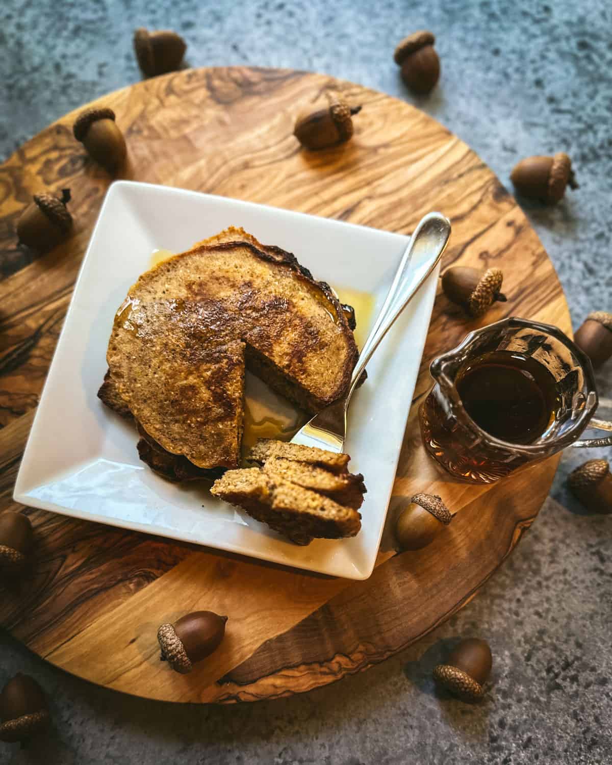 An acorn pancake with a bite cut, on a square white plate with syrup and butter. On a circular wood cutting board surrounded by acorns and a small dish of maple syrup. 