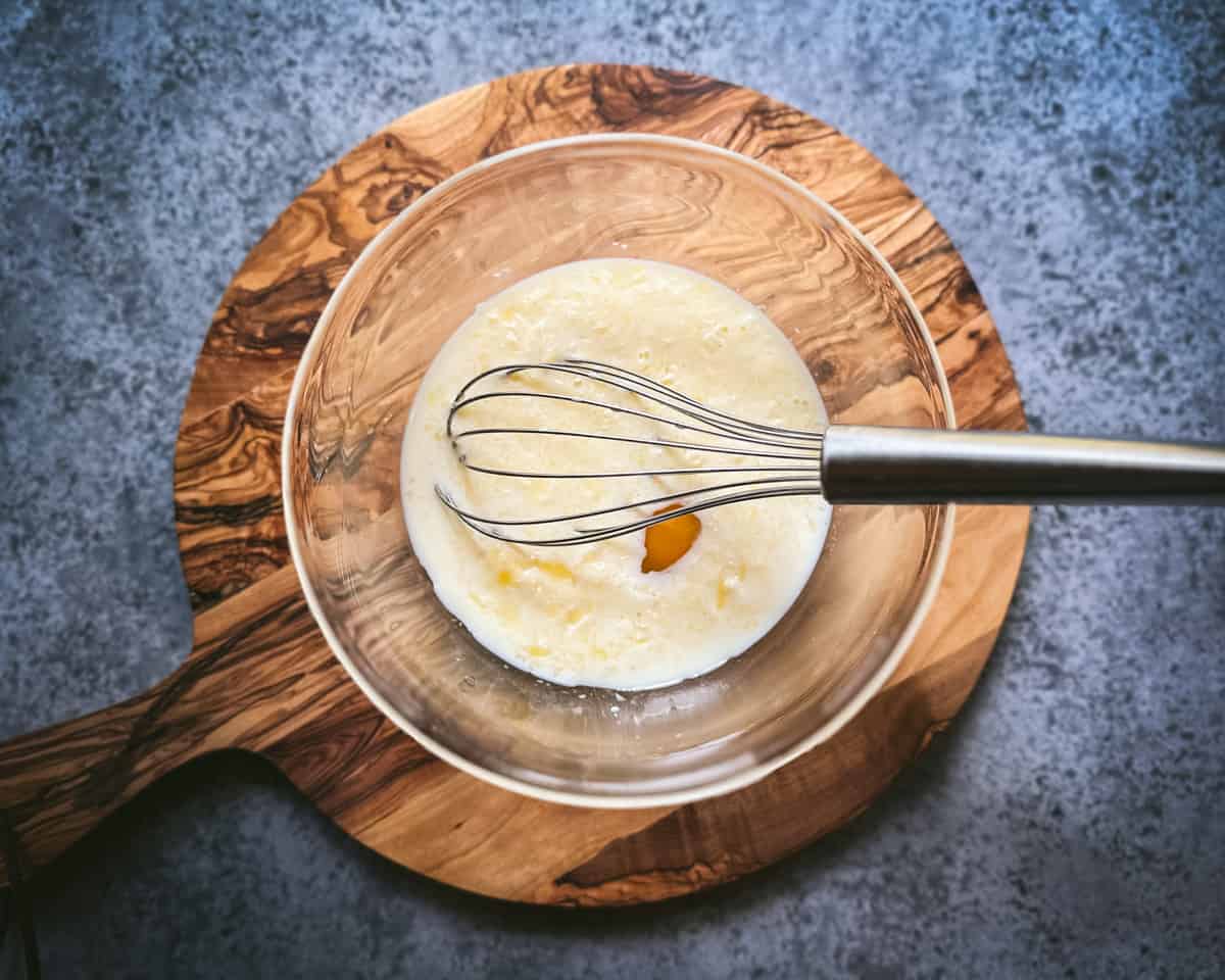 A whisk in the wet ingredients in a bowl on a circular wood cutting board on a gray countertop. 