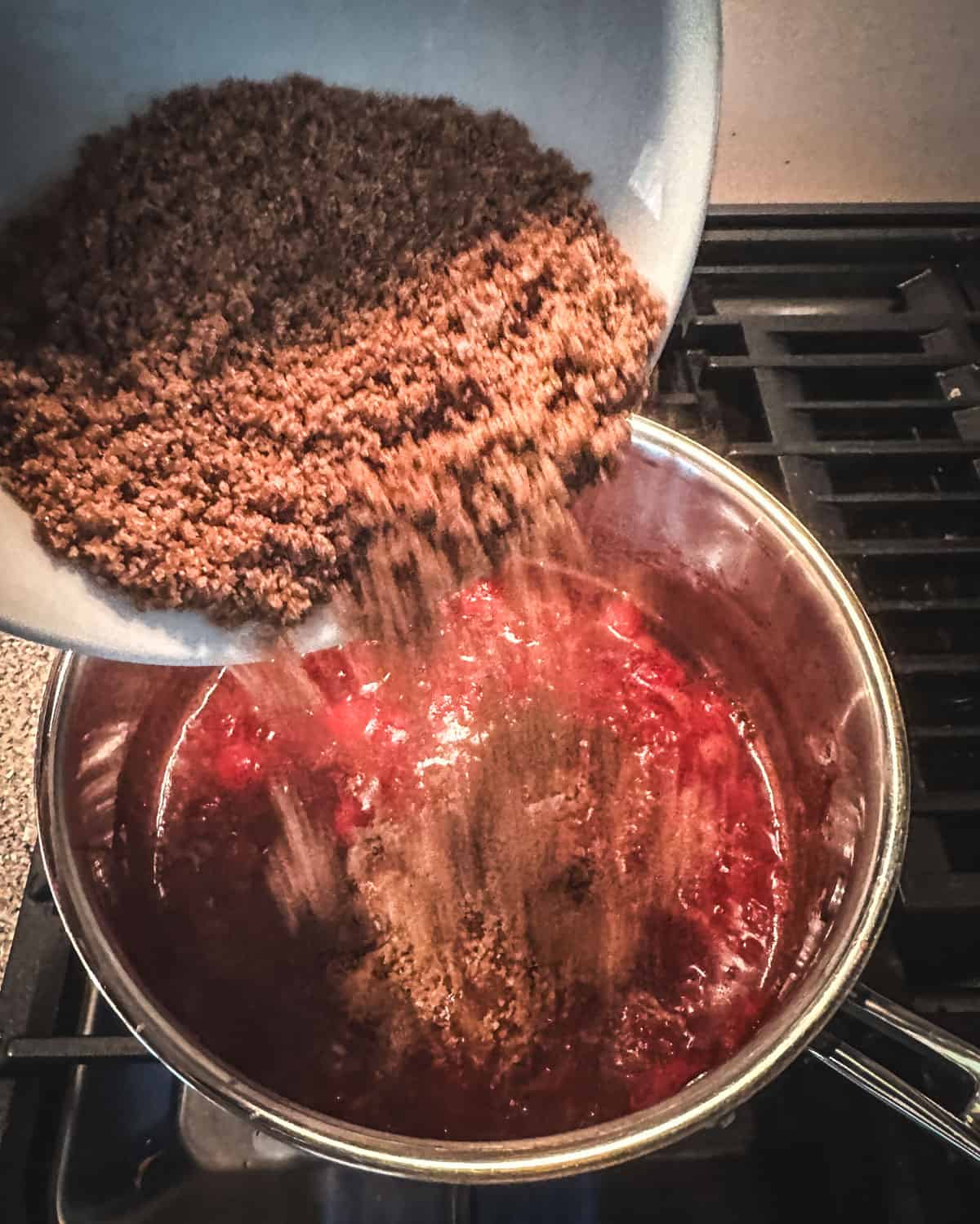 Brown sugar and pectin mix pouring from a white bowl into a pot with the boiled cranberries and spices. 