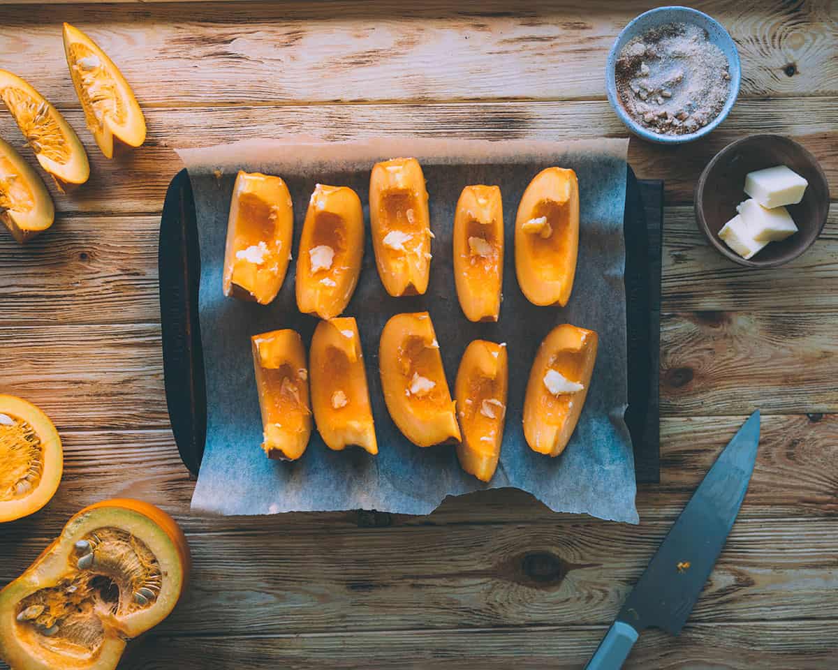 Pumpkin wedges skin side down sitting on a parchment paper lined cookie sheet, on a wood surface surrounded by ingredients. 