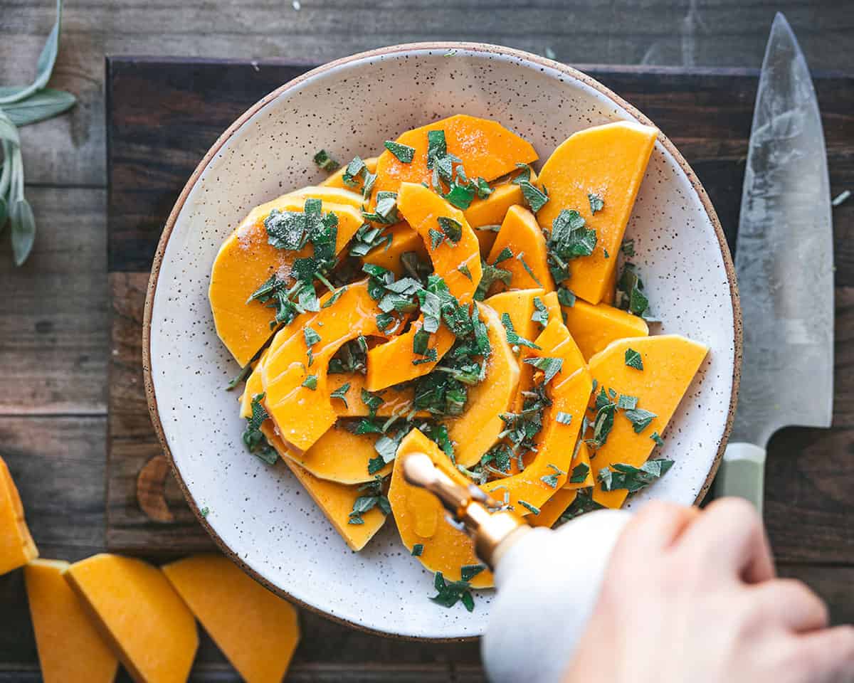 A white bowl with butternut squash slices tossed with fresh sage and olive oil being drizzled on, on a dark wood surface. Top view. 