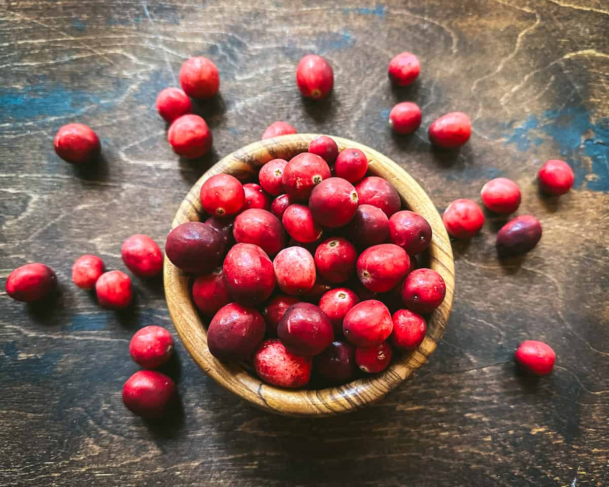 A wood bowl full of fresh cranberries, on a dark wood surface surrounded by loose fresh cranberries. Top view.