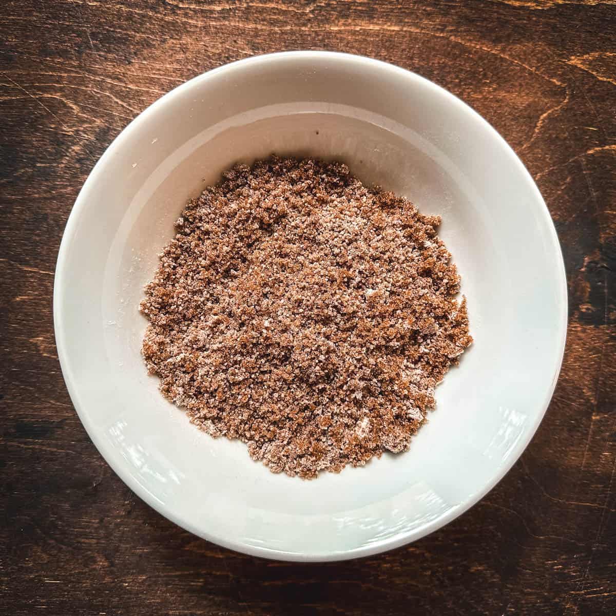 Brown sugar and pectin powder in a small white bowl sitting on a dark wood surface, top view. 