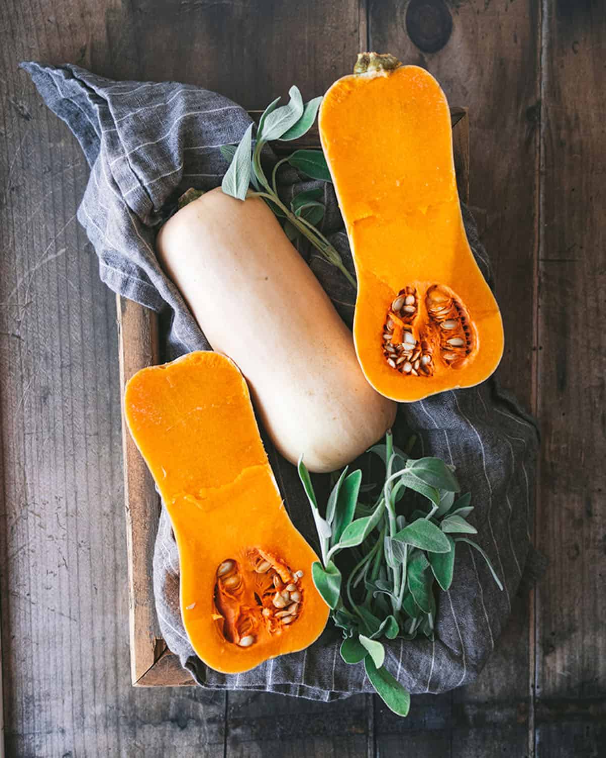 2 halves of butternut squash face up with a whole butternut squash in the middle, on a dark wood surface with a grey towel, surrounded by fresh sage. 