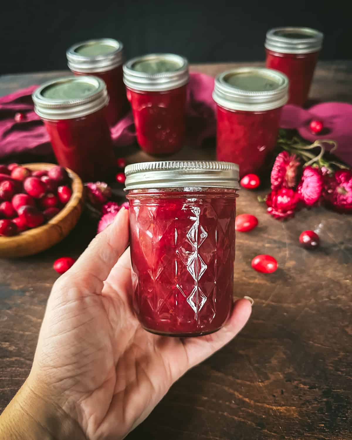 A jar of canned cranberry sauce being held by a hand, with other jars of cranberry sauce, fresh cranberries, and a burgundy cloth in the background.