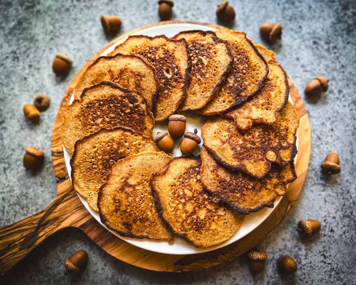 A plate with acorn pancakes layered in a circle, on a round wood cutting board, on a gray countertop surrounded by acorns.