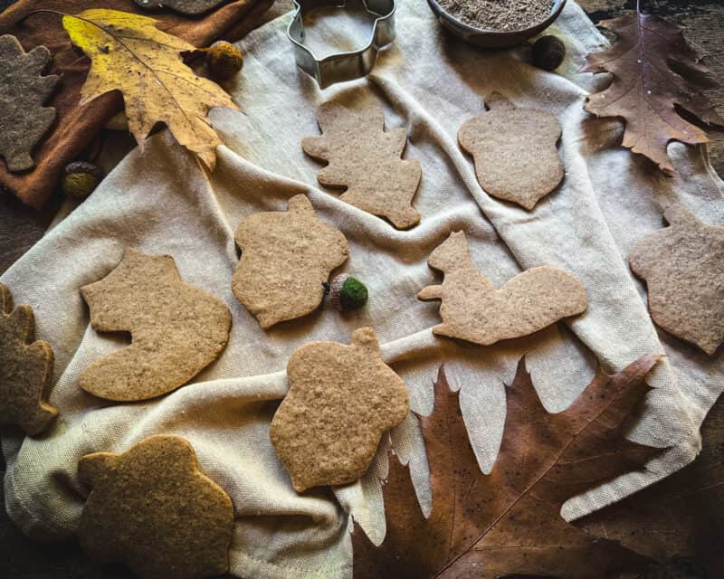 Acorn flour cookies in fall shapes on a white fabric, surrounded by an acorn cookie cutter, fall leaves, and a bowl of acorn flower.