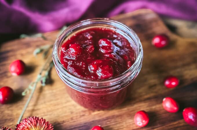 A jar of cranberry sauce opened, on a dark wood surface surrounded by fresh cranberries, and dried flowers.