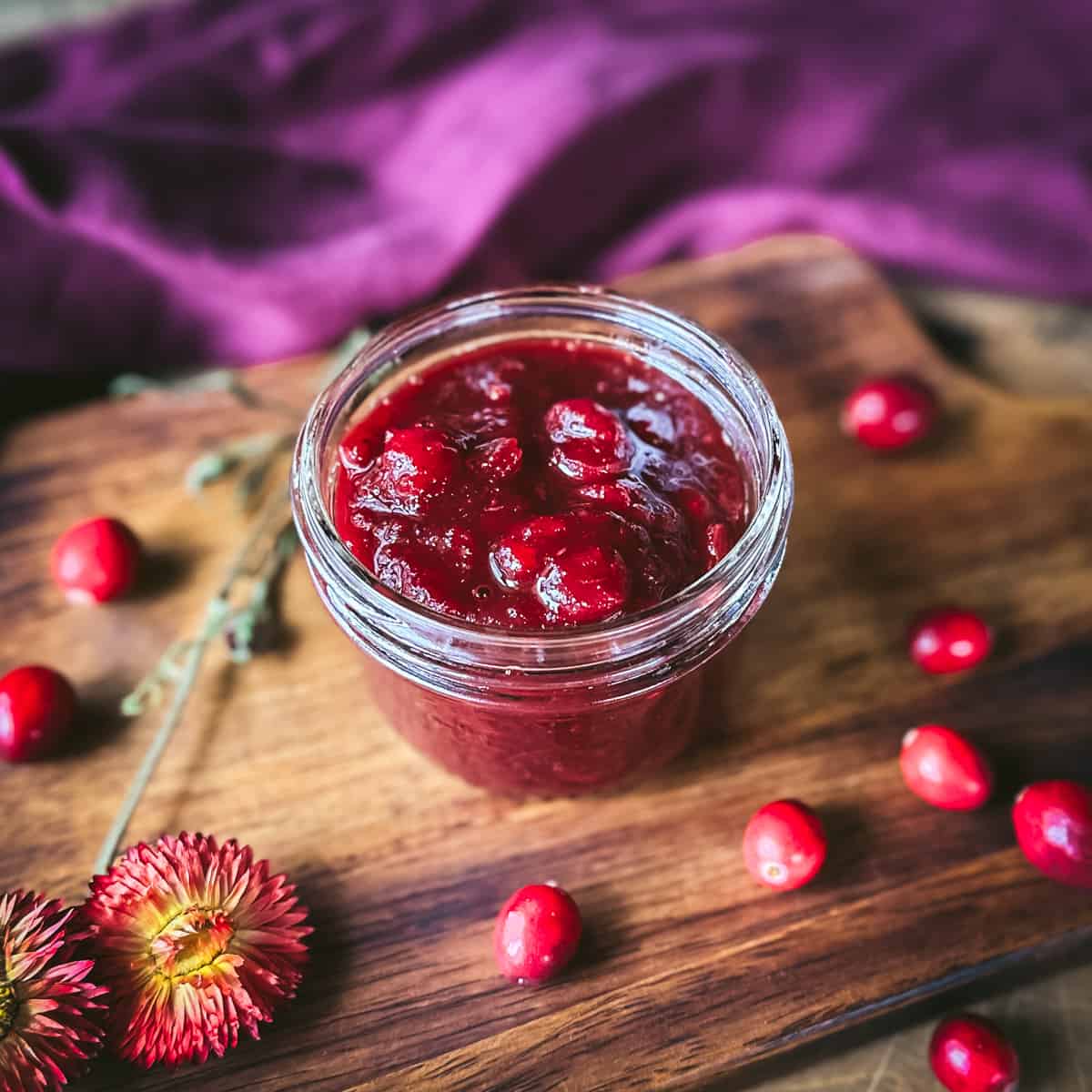A jar of cranberry sauce opened, on a dark wood surface surrounded by fresh cranberries, and dried flowers.