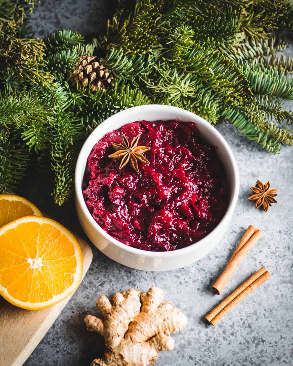 A white bowl of cranberry sauce with a star anise in it, on a counter surrounded by orange slices, cinnamon sticks, ginger, and evergreen branches. 