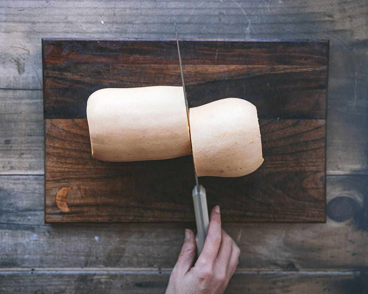 A butternut squash cut in half crosswise with a hand on the knife, on a dark wood cutting board. 