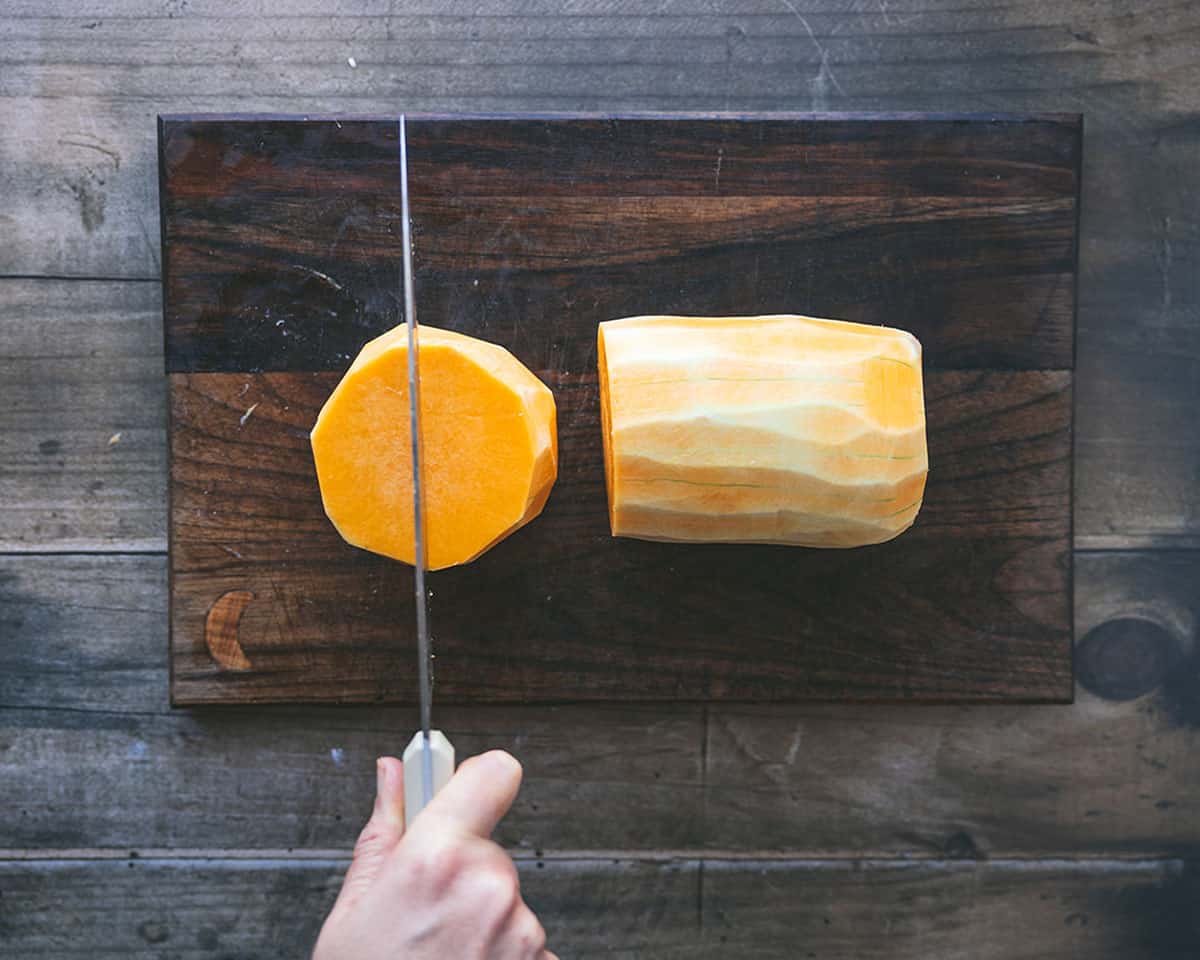 Half of a butternut squash standing upright being sliced in half with a knife, with the other half laying on its side. On a dark wood cutting board. 