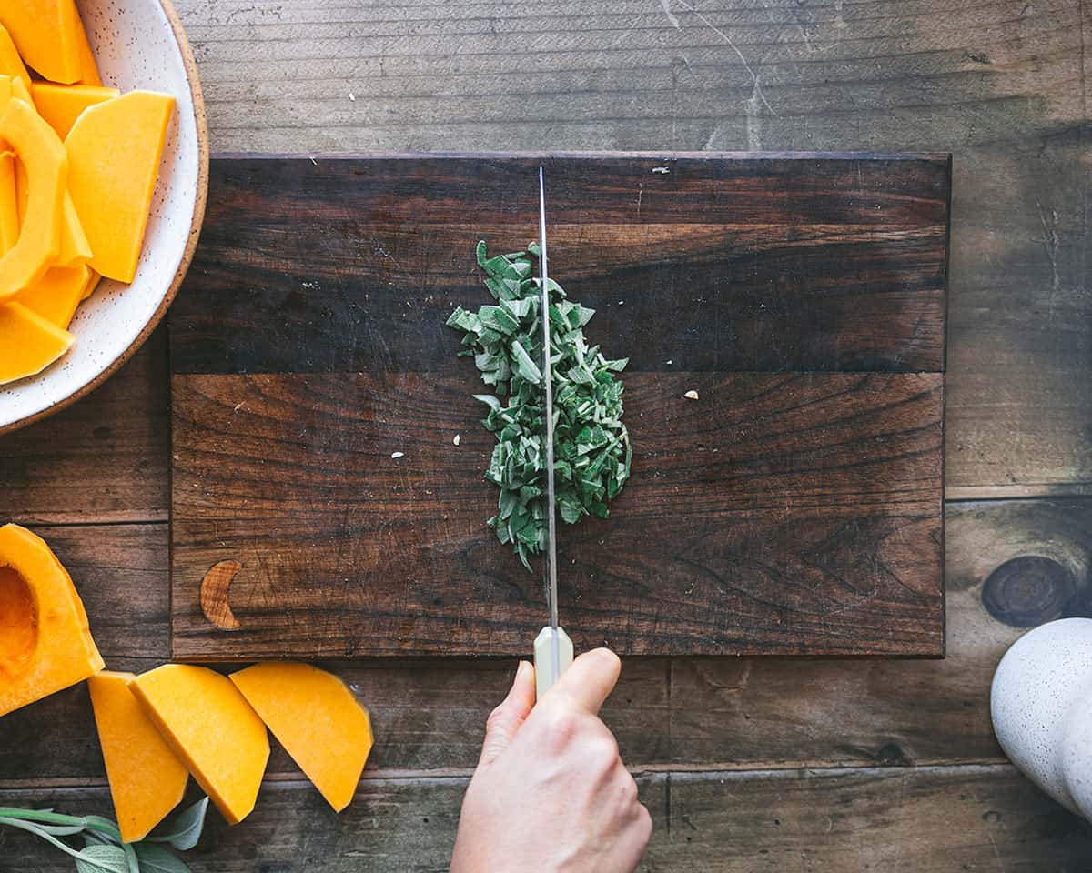 Fresh sage being chopped with a knife on a dark wood cutting board, surrounded by half-moons of butternut squash, and a bowl of slices. 