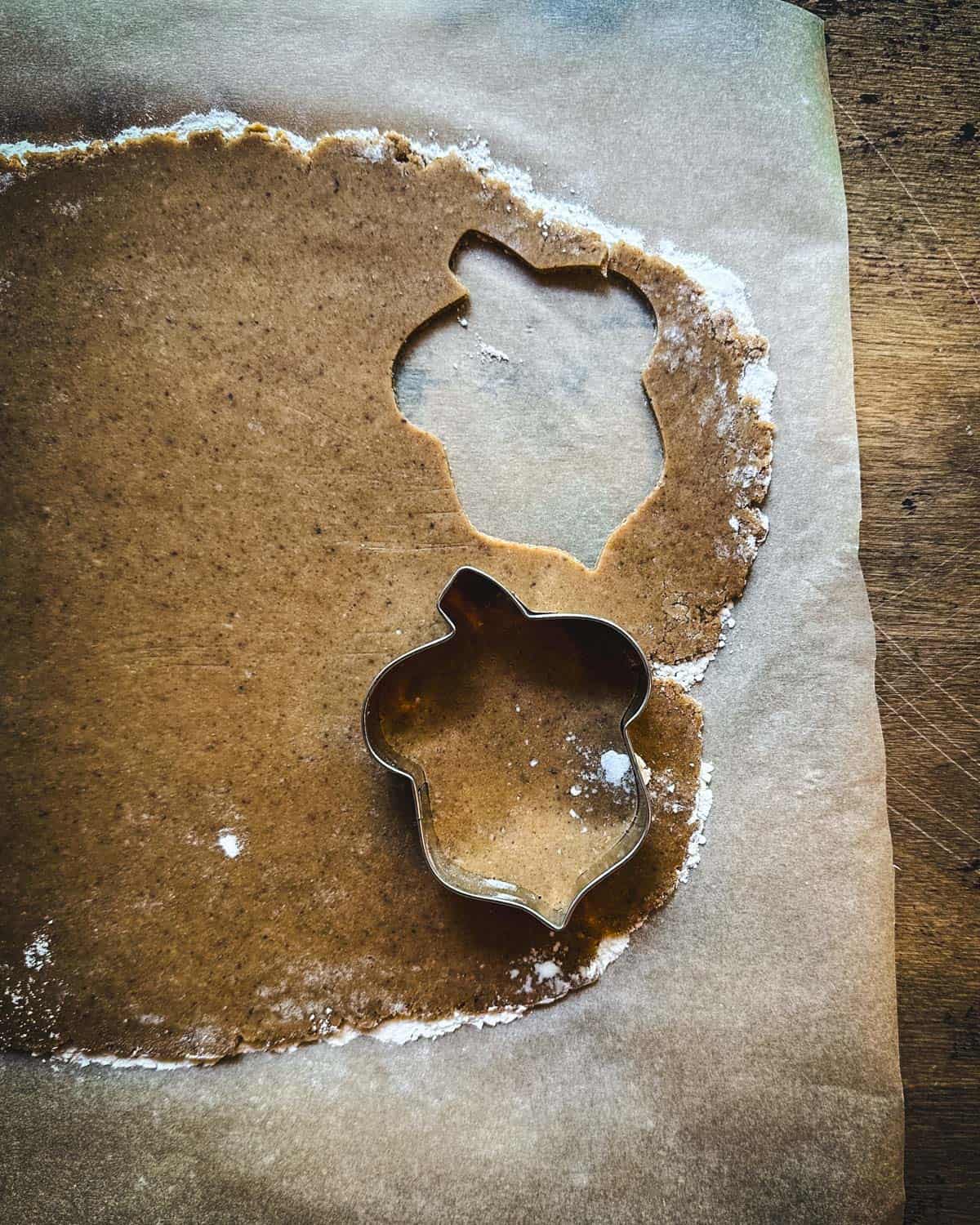 A rolled out dough with an acorn shaped cookie cutter on it, and an acorn shaped cookie cut out of it already. 