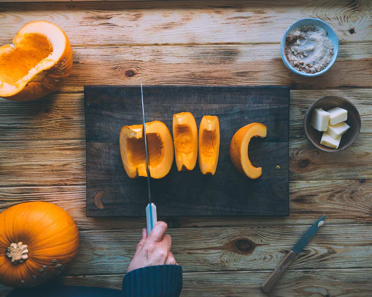 A sugar pumpkin being cut on a wood cutting board, sitting on a lighter wood surface, surrounded by pumpkin roasting ingredients. 