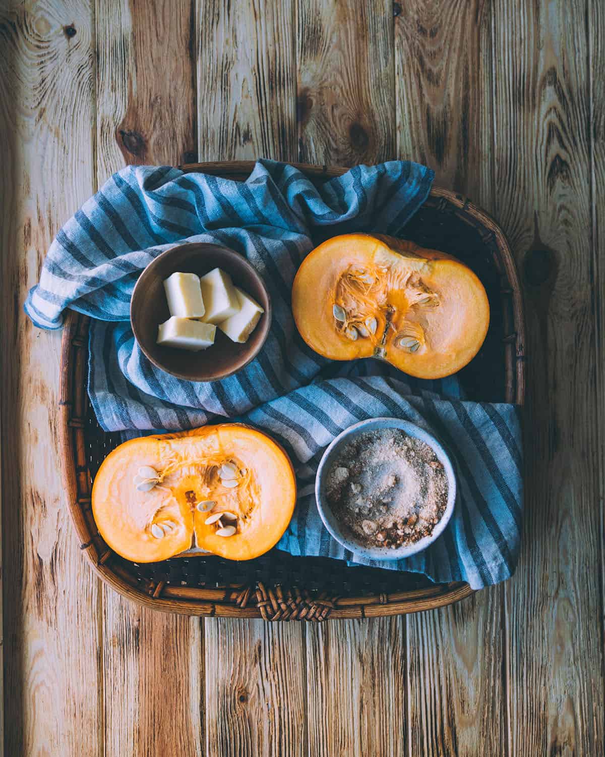 A sugar pumpkin cut in half, face up with butter and sugar with cinnamon in a bowl and butter in another small bowl, on a tray with a blue striped cloth, sitting on a wood surface. 