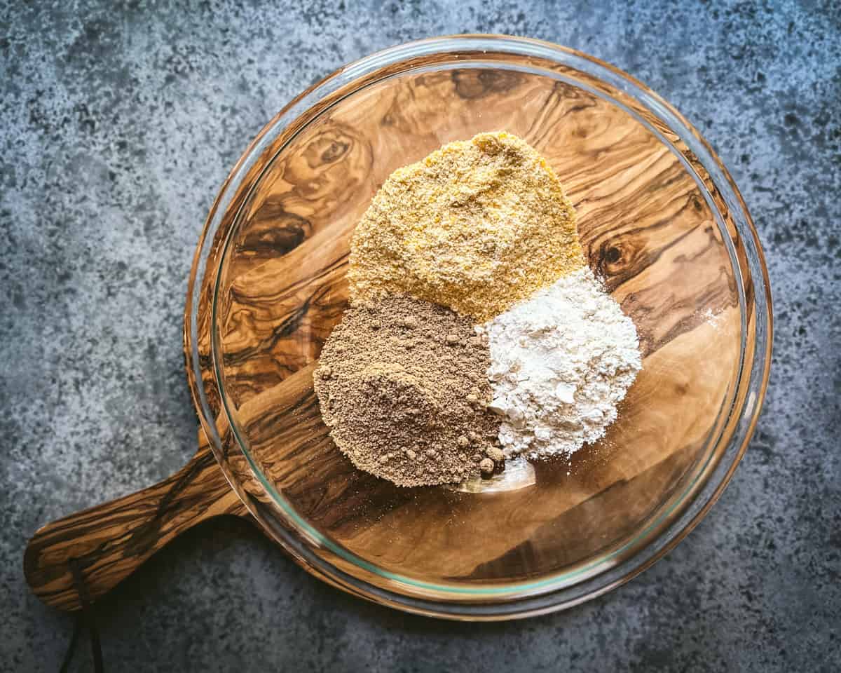 3 flours piled in a medium glass mixing bowl on a circular wood cutting board. 