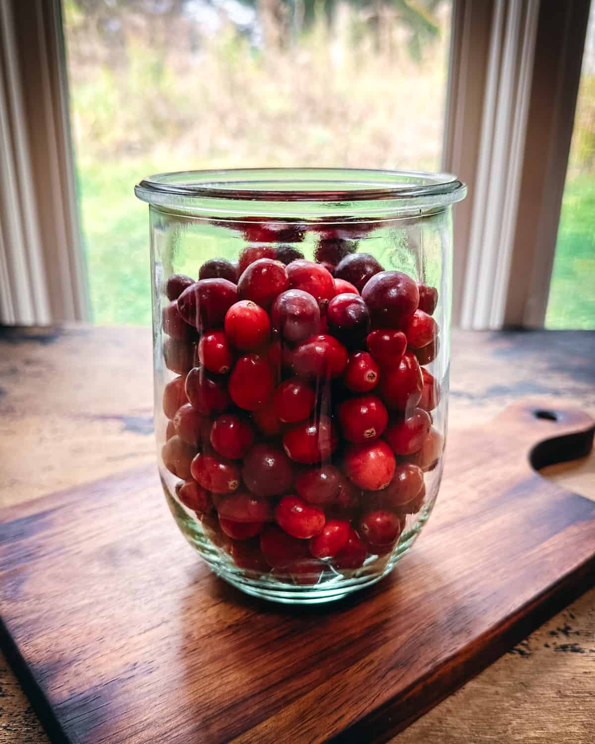A jar of fresh cranberries sitting on a wood cutting board, with a window in the background.