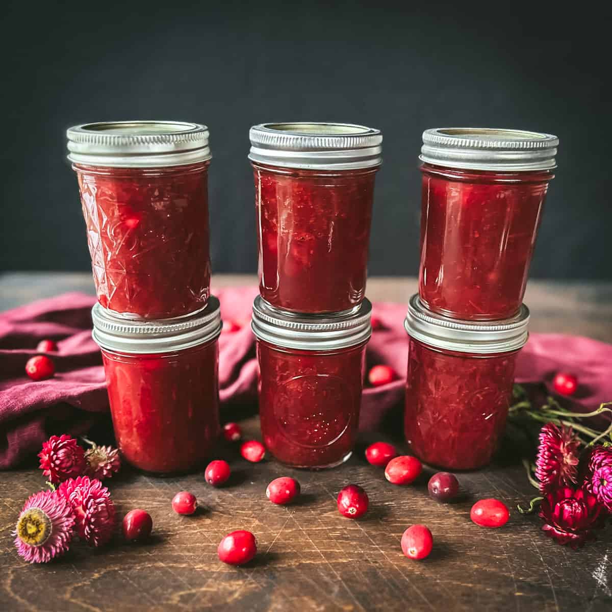 6 jars of canned cranberry stacked 2 high, on a dark wood surface, surrounded by fresh cranberries, dried flowers, and a burgundy cloth. 