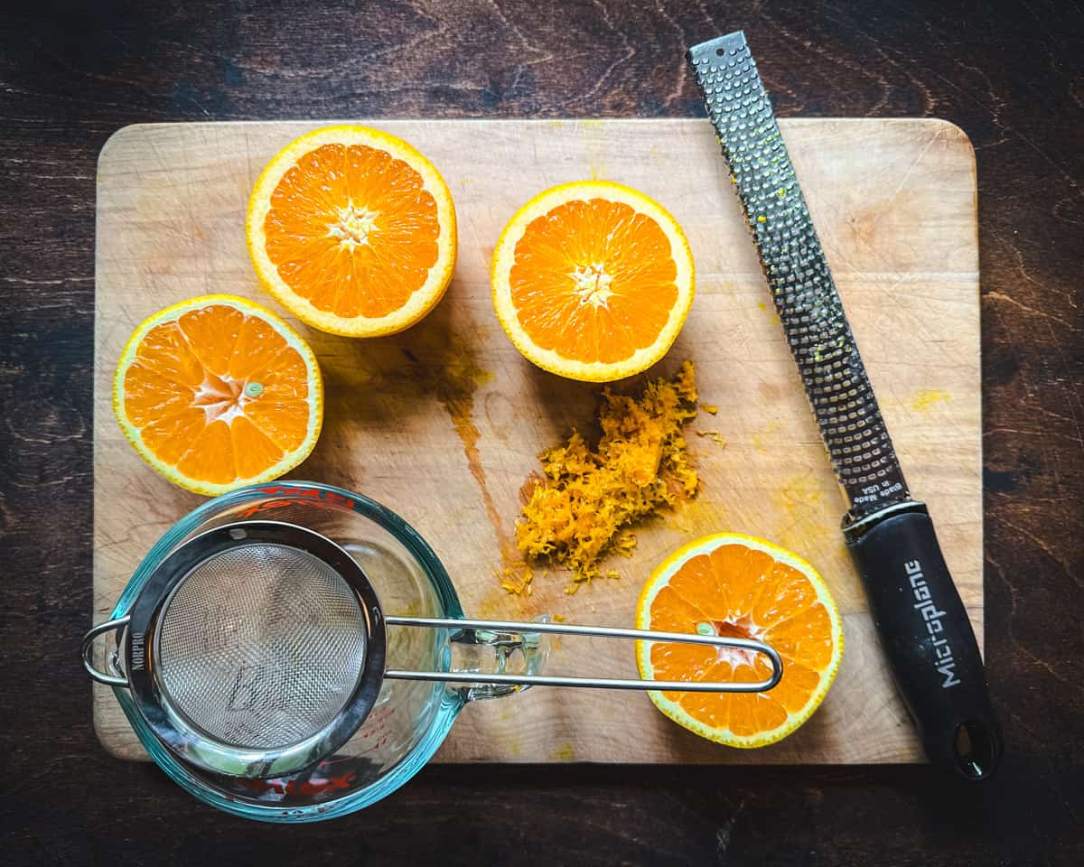 2 oranges cut in half, face up on a wood cutting board, with zest. On the cutting board is a small strainer on a small bowl and a microplane. 