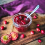 An open jar of pickled cranberries with a spoon in it, on a wood cutting board surrounded by fresh cranberries, dried flowers, and a burgundy cloth.