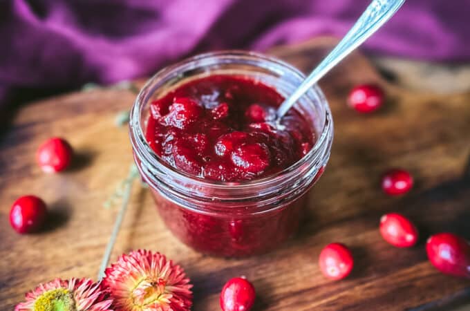 An open jar of pickled cranberries with a spoon in it, on a wood cutting board surrounded by fresh cranberries, dried flowers, and a burgundy cloth.