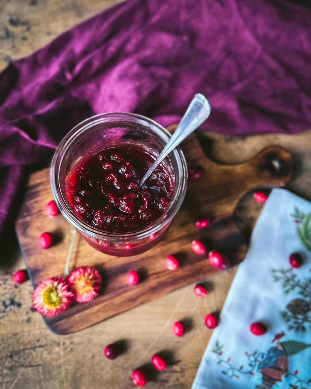A jar of pickled cranberries with a spoon in it, on a wood surface surrounded by fresh cranberries, dried flowers, and a burgundy cloth. 