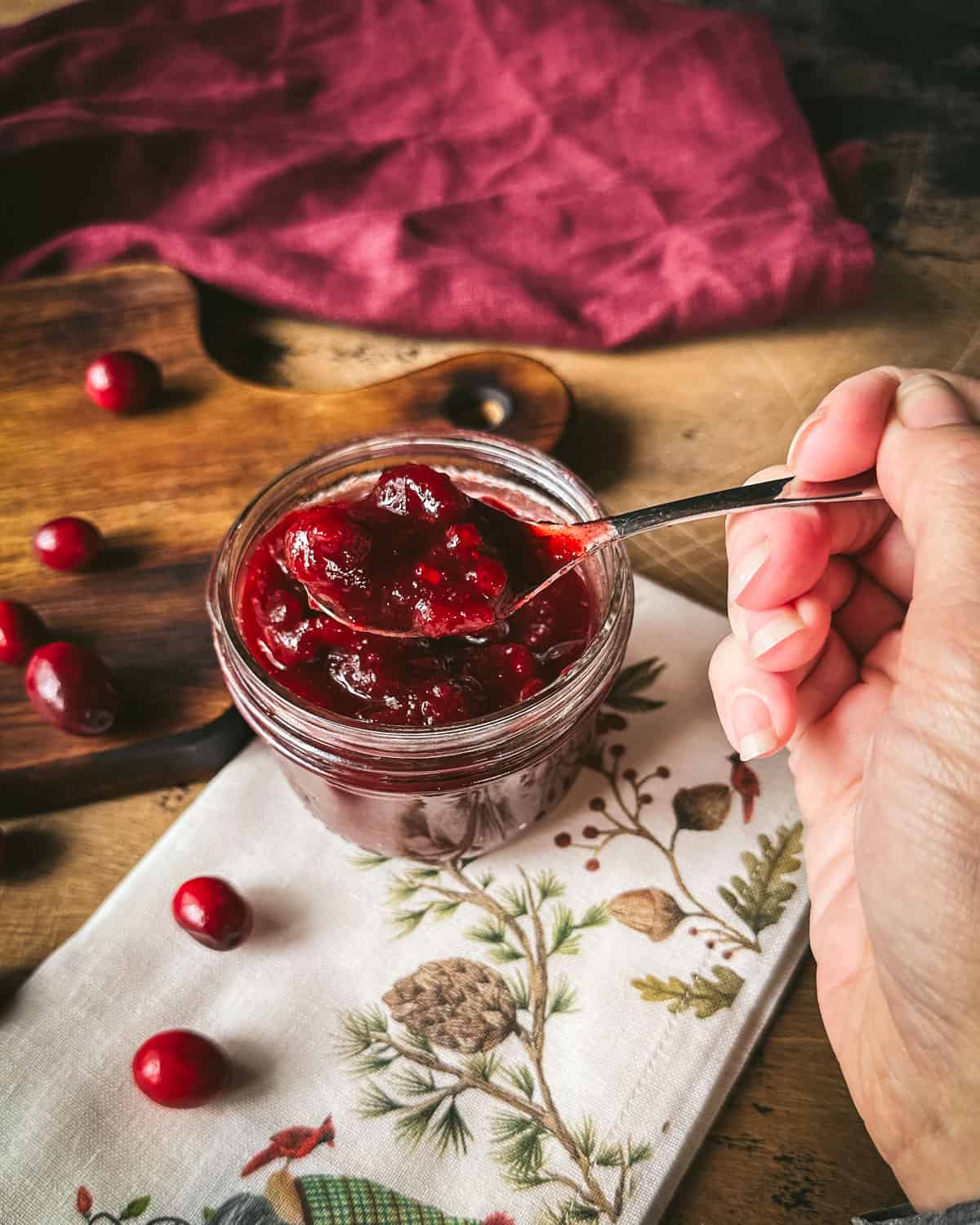Pickled cranberries being spooned out of a jar, sitting on a white napkin with flowers, on a wood cutting board, surrounded by fresh cranberries.
