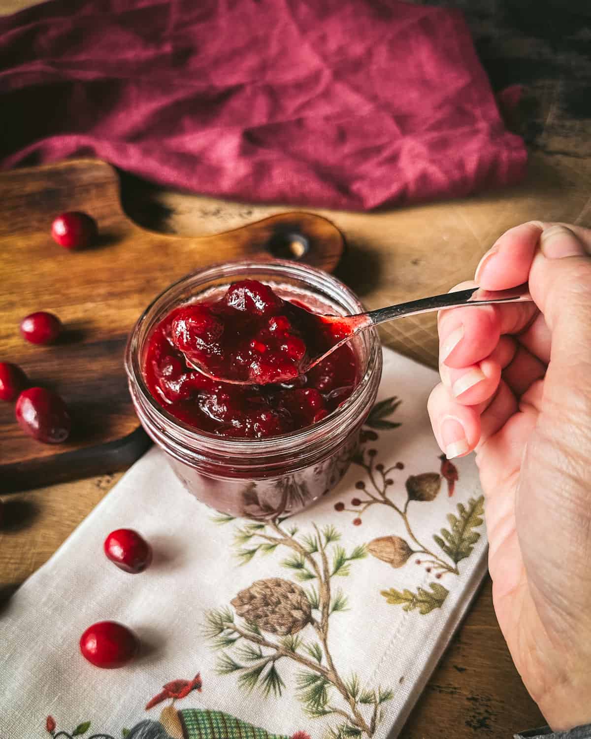 Pickled cranberries being spooned out of a jar, sitting on a white napkin with flowers, on a wood cutting board, surrounded by fresh cranberries. 