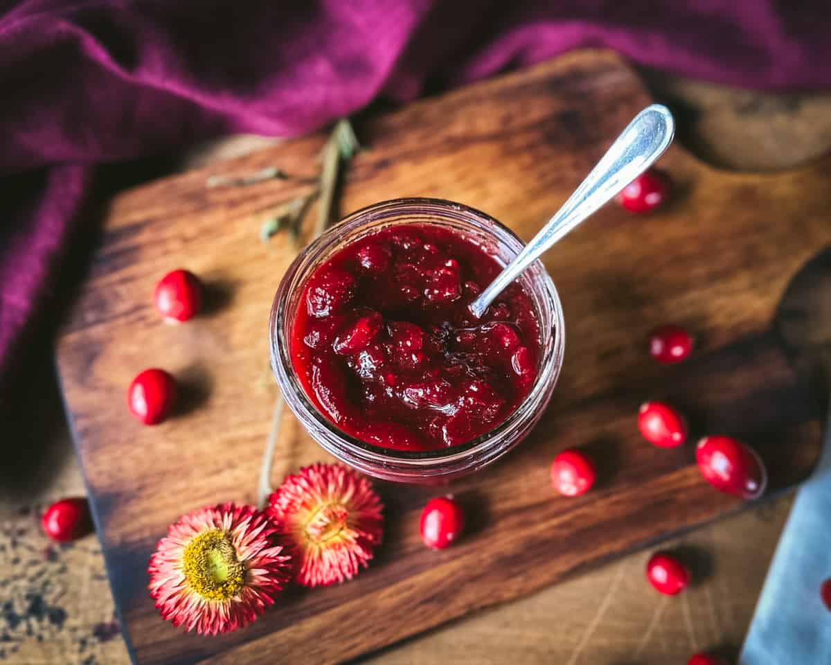 A jar of pickled cranberries with a spoon in it, on a wood surface, with fresh cranberries, dried flowers, and a burgundy cloth surrounding.